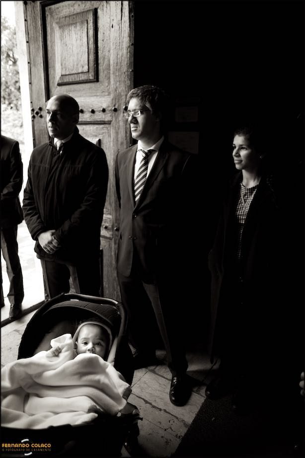 Bebé, dentro do seu carrinho, junto da mãe e do pai à porta da igreja.