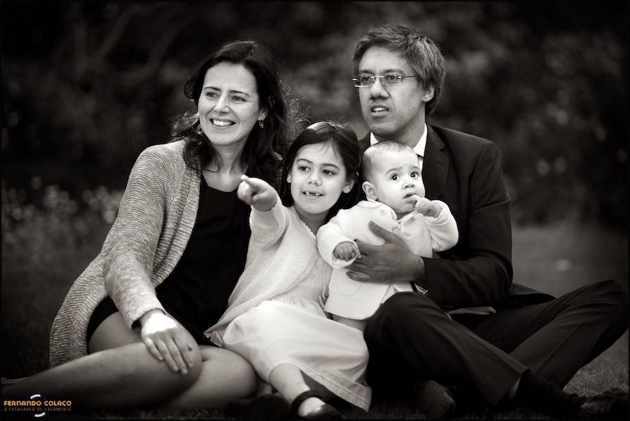 Família do bebé que foi batizado, pais e irmão, sentados no chão num retrato no jardim.