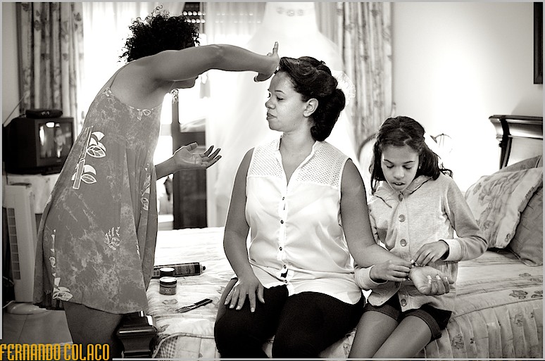 A noiva sentada numa cama, com a filha a pôr-lhe a pulseira no pulso e uma amiga a retocar o penteado.