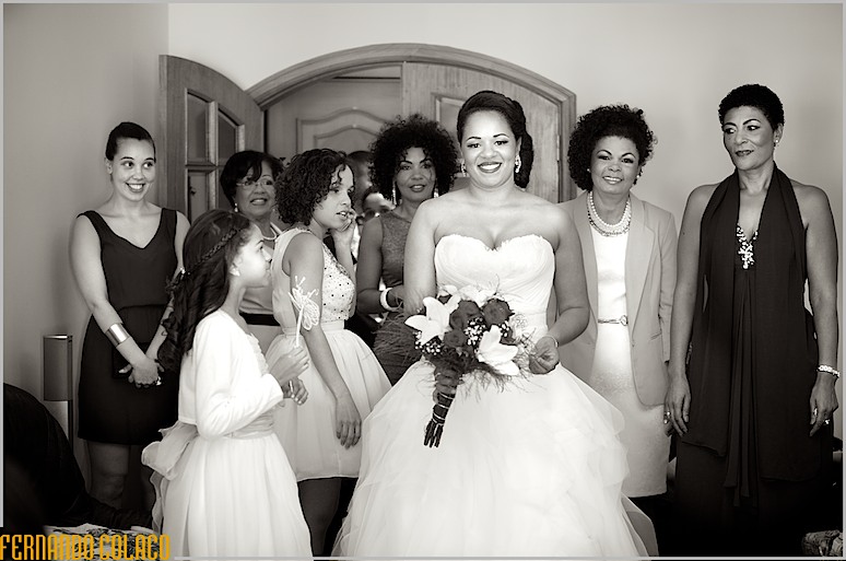 A noiva em frente de todas as mulheres presentes, quando da sua preparação, com o ramo de flores na mão.