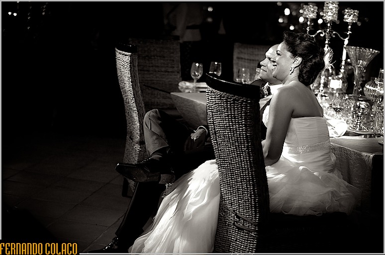 Os noivos, sentados, por entre duas cadeiras olham satisfeitos para algo em frente deles durante a festa do casamento na Quinta do Roseiral.