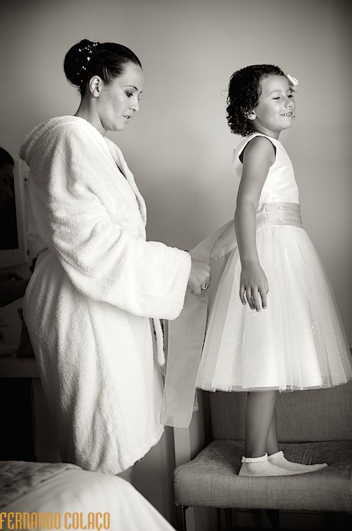 A noiva e a filha, sobre uma cadeira, enquanto lhe aperta um grande laço na cintura do vestido.