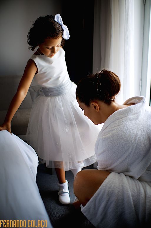 A filha da noiva, perto de uma janela, com a mãe, que lhe calça os sapatos.