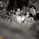 Por entre os tufos de folhas das árvores do jardim Quinta de D. Nuno em Fátima, os noivos conversam, com a filha no meio deles, vistos pelo fotógrafo de casamento em Leiria.