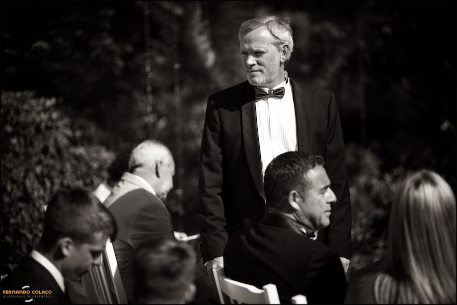 The groom, standing with seated guests, waits for the bride to arrive for the wedding ceremony.
