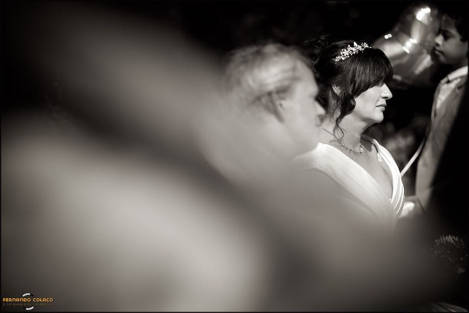 The bride, in profile, seen among unfocused guests at the wedding ceremony.