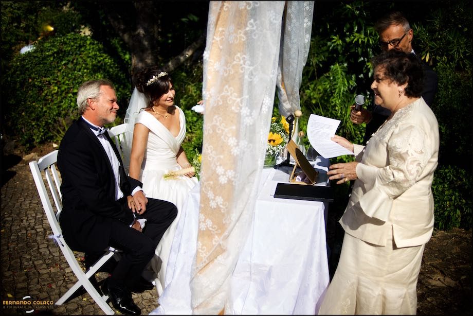 The bride and groom, seated at the ceremonial table, listen to the mother of the bride, in a speech, next to the conservative officiant of the ceremony.