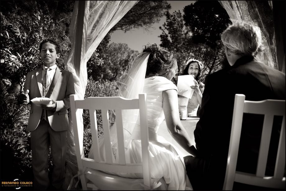 The bride and groom, with their backs turned, next to their youngest son with the wedding ring pillow in hand, listening to a wedding guest, in a speech during the wedding ceremony.