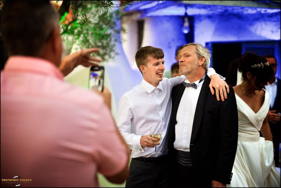 The groom with a wedding guest who hugs him, while another takes a photograph.