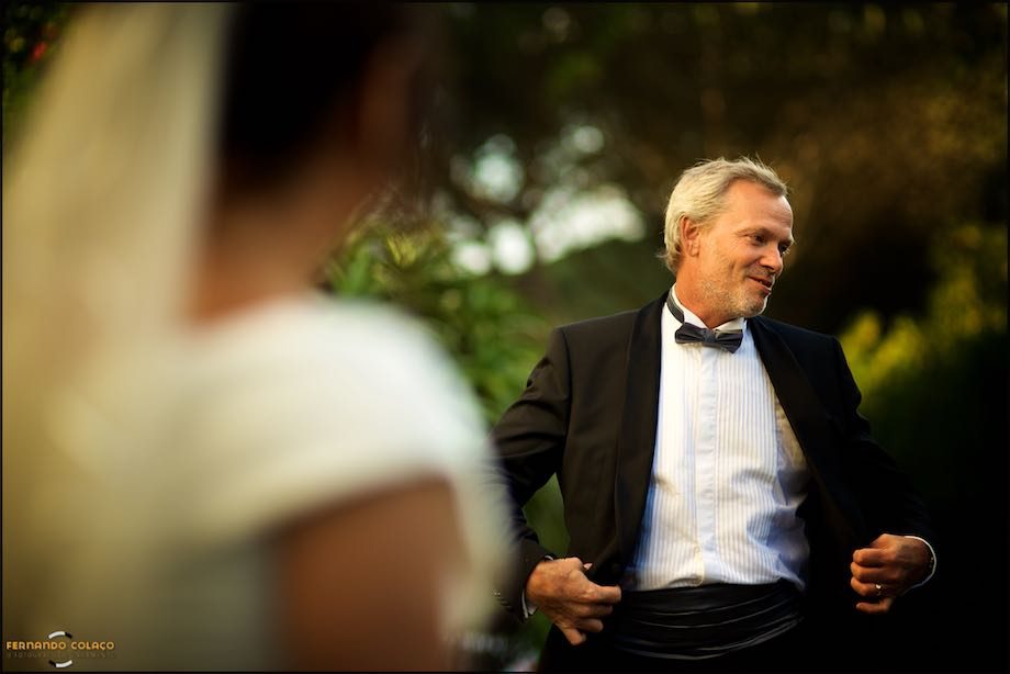 With the bride, out of focus, in front, the groom looks to the side with hands on his coat.
