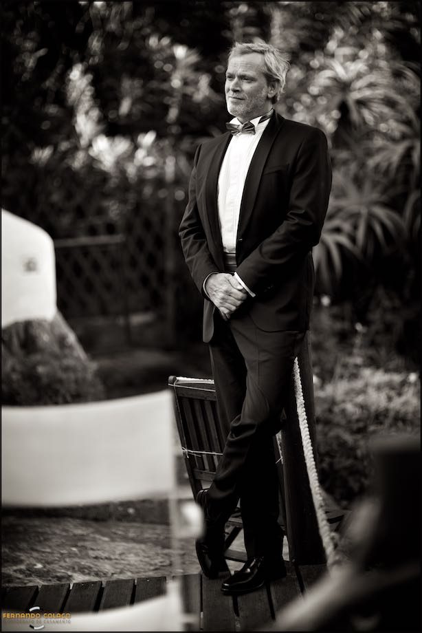 The groom, standing and leaning against a pole in the garden of Morada do Sol in Sintra, hands together, looks to the side.