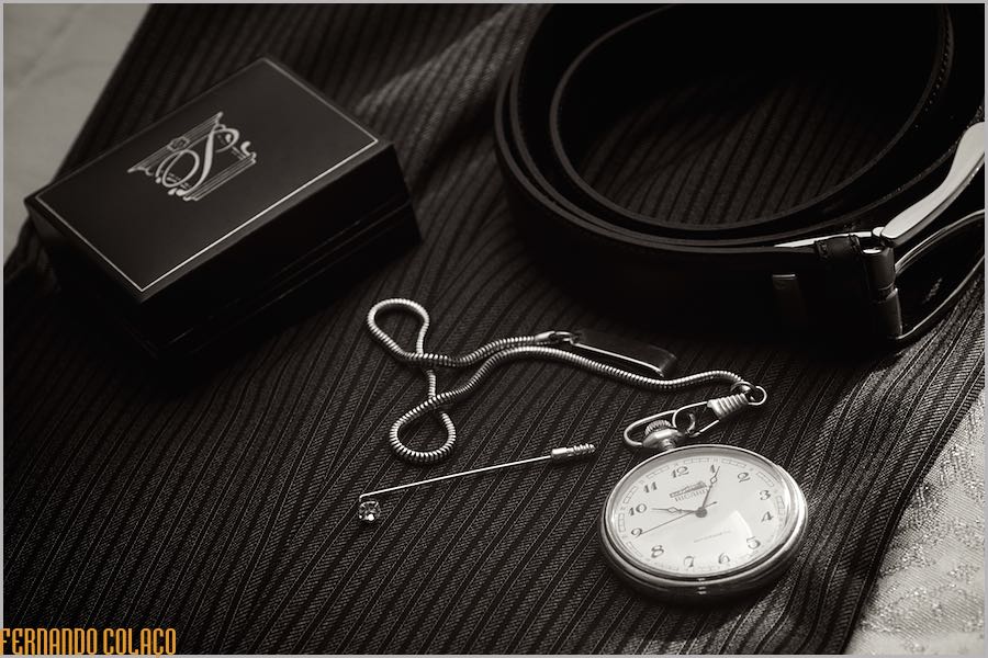 O relógio de bolso que o noivo vai usar no dia do casamento, junto do cinto e de uma caixa, sobre o tecido das calças.