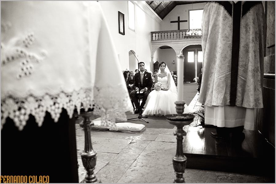 Vistos por entre o padre, na sua homilia, e parte da altar, os noivos, ao fundo, sentados na frente dos seus convidados.