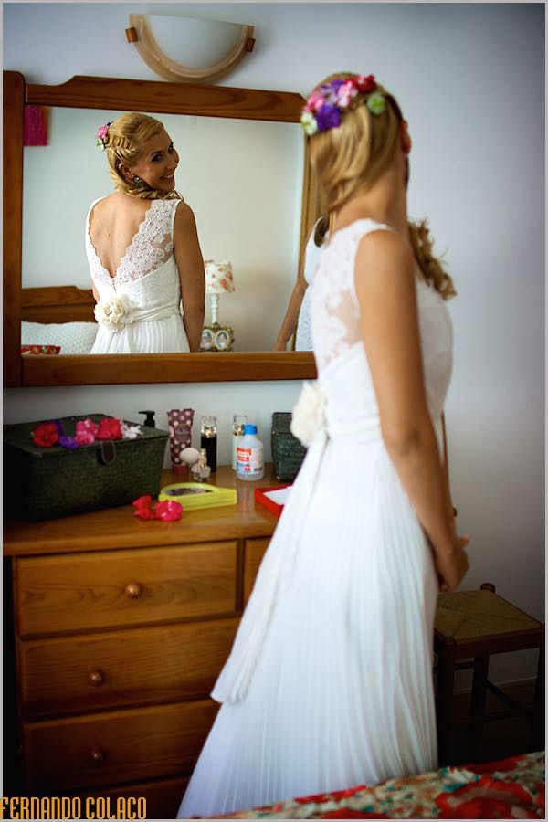 Standing by a mirror, the bride looks into it to see the back of her dress.