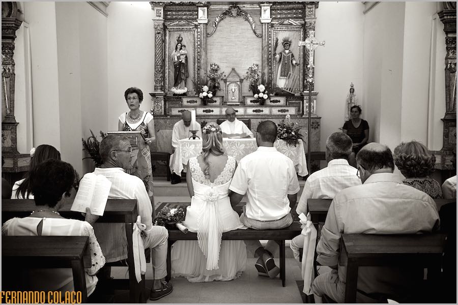 Seen from behind, the bride and groom, seated, and some wedding guests, while a guest reads the sacred texts.