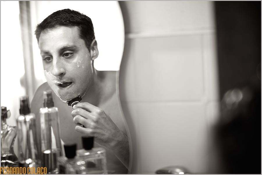 Seen in a mirror and with several bottles in front of him, the groom shaves his beard to prepare for the wedding.