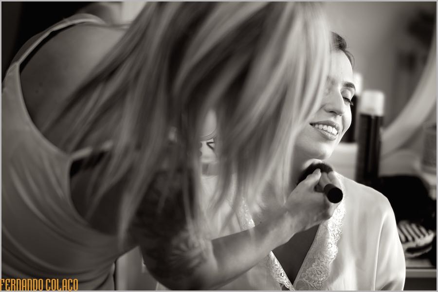 Looking to the side with her eyes closed, the bride smiles as the makeup artist, blurred in the foreground, smoothes her makeup with a brush.