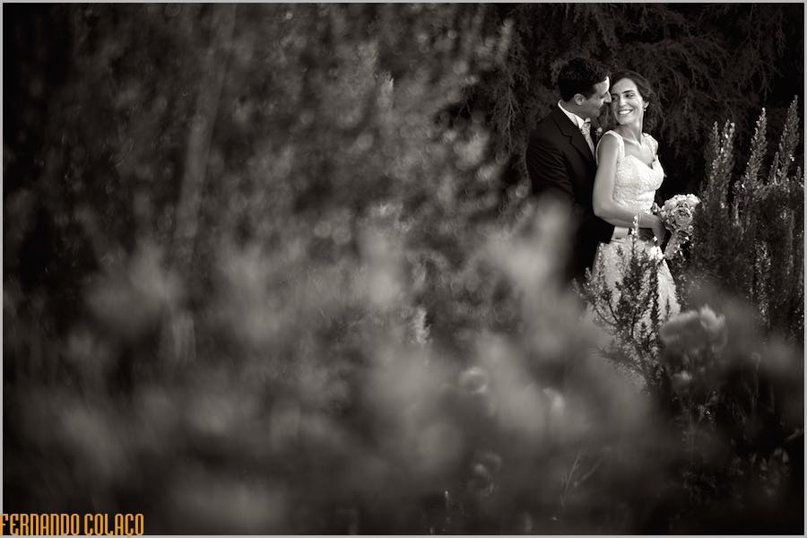 Leaning on the groom's chest and cheeks together, the bride looks back smiling, in the midst of wild, unfocused bushes in the garden.