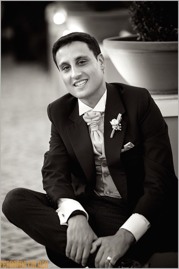 The groom smiles, sitting by a wall with vases of flowers.