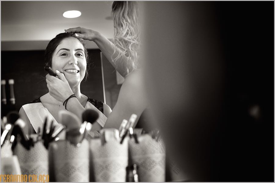 In a mirror at the hairdresser's, the makeup artist, with her back turned and out of focus, applies powder with a brush to the bride's face as she prepares for the wedding.