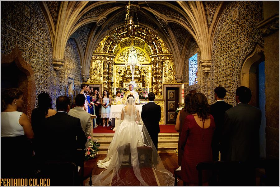 Interior of the church, with walls full of blue tiles, Manueline arches and gilded carvings on the altar, with the bride and groom and guests seen from behind.