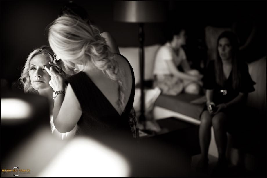 Detalhes da maquilhagem da noiva com amigas sentadas ao fundo, desfocadas.