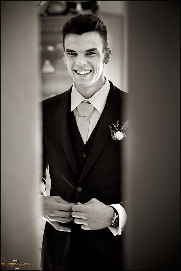 Depois de pronto para ir para a cerimónia do casamento, o noivo abotoa os botões do casaco, espreitado na abertura de uma porta pelo fotógrafo de casamento.