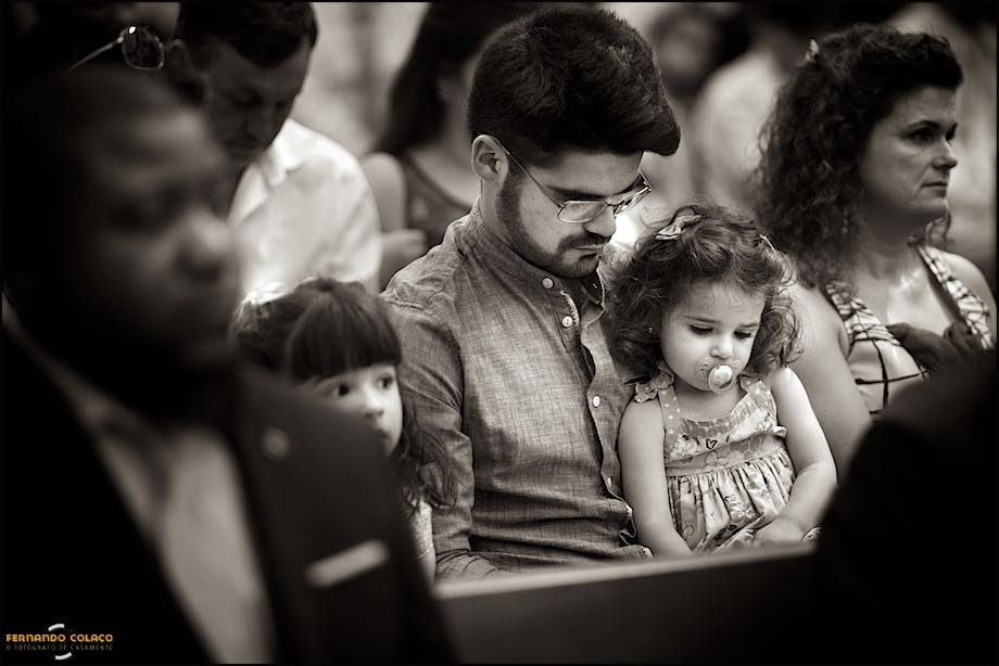 No meio de outros convidados do casamento na Igreja Paroquial de Nossa Senhora da Assunção em Cascais, um pai, com a sua filha pequenina ao colo, lê com ela algo num livro.