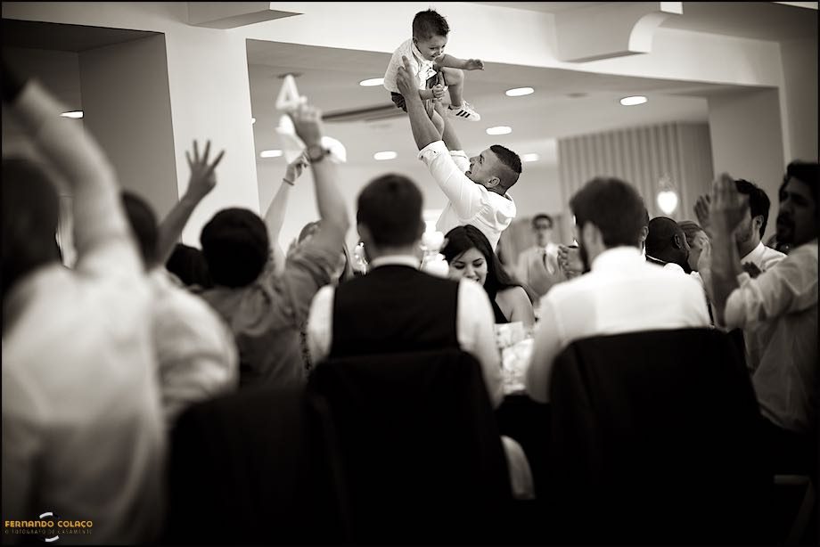 No meio de outros convidados do casamento à mesa da refeição, um deles eleva o seu filho pequeno no ar, numa brincadeira com ele.