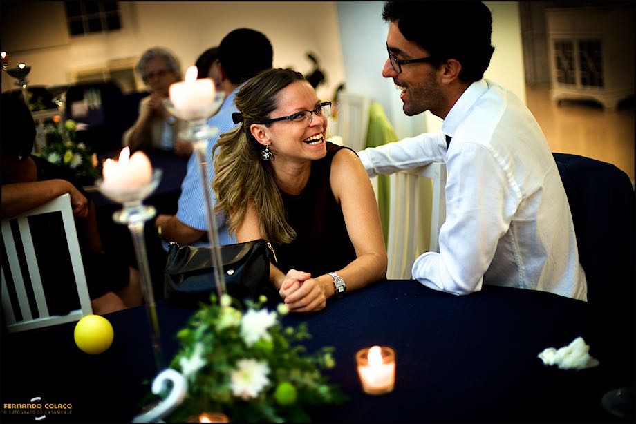 Sentados numa mesa durante a fresta do casamento, um casal ri-se olhando-se de frente.