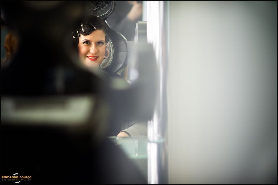 No cabeleireiro, a face da noiva, sorrindo, vista num pequeno espelho redondo.