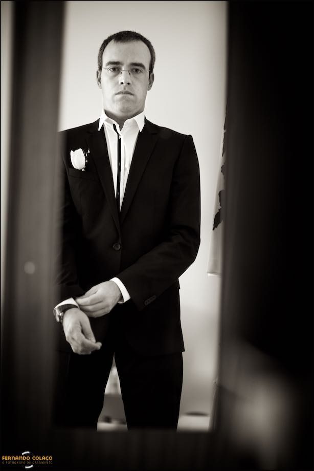 O noivo a ver-se ao espelho de móvel da casa, acertando o fato do casamento.