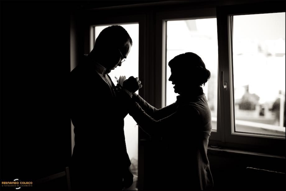 Em silhueta junto de uma janela, a noiva põe o pequeno ramo de flores na lapela do casaco do noivo, para o casamento.