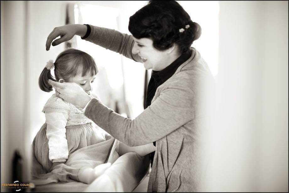 A mãe, e noiva, penteado a sua pequena filha, acabada de vestir sobre um móvel da casa.