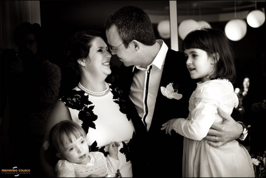 Os noivos, depois da cerimónia de casamento, de faces muito juntas olhando um para o outro, cada um com uma filha ao colo.