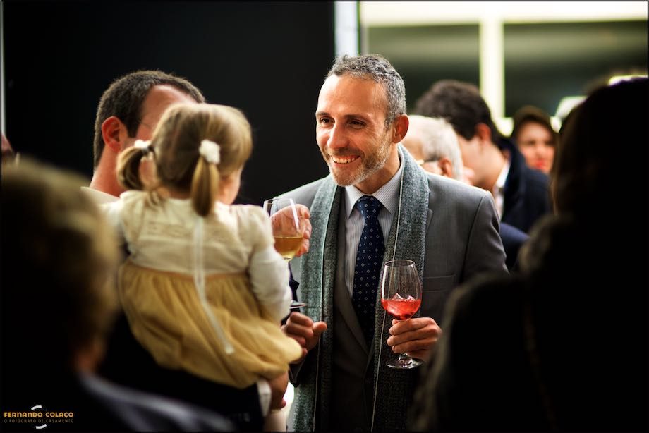 Um convidado do casamento, no convívio depois da cerimónia, conversando com a filha mais nova do casal recém casado, ao colo do pai, o noivo.