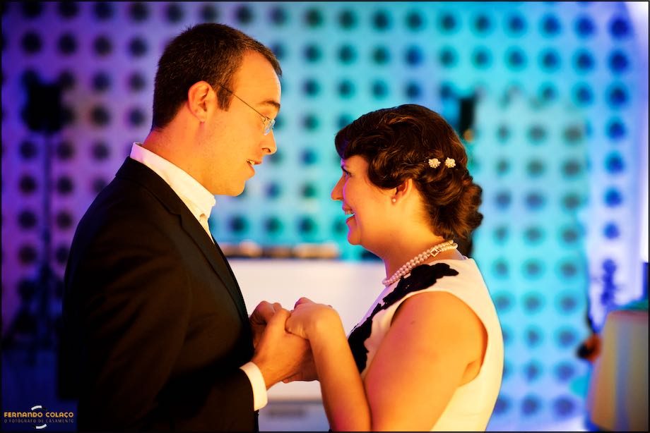 Pintados de luz laranja sobre um fundo azul, os noivos de frente um para o outro dão as mãos, durante a festa do casamento.