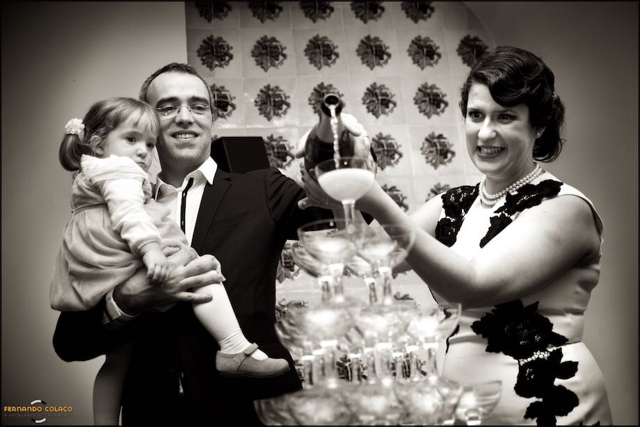Os noivos despejam uma garrafa de champanhe sobre uma pirâmide de copos, com a filha mais nova ao colo do noivo.