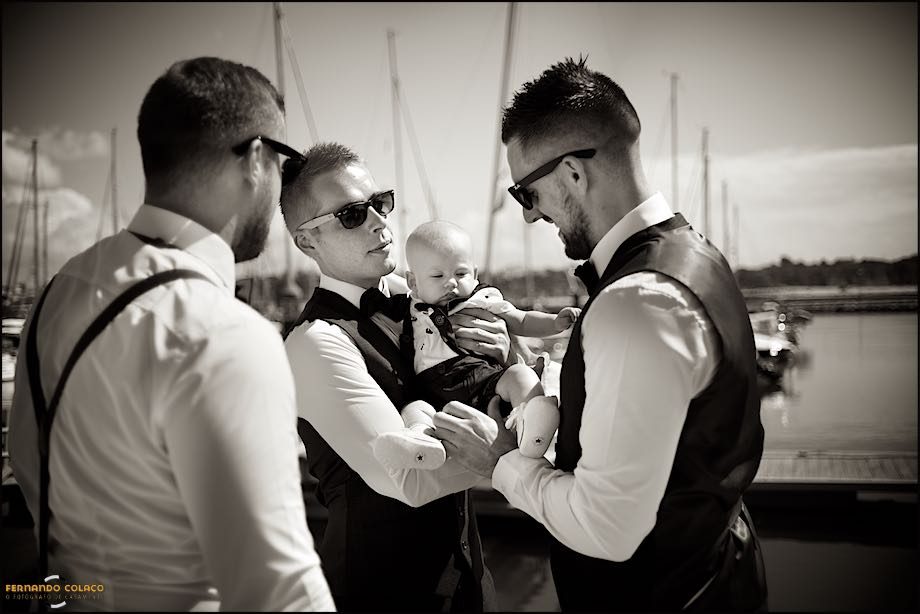 Next to a marina in the Algarve, the groom caresses his baby son, on the lap of one of the wedding guests.