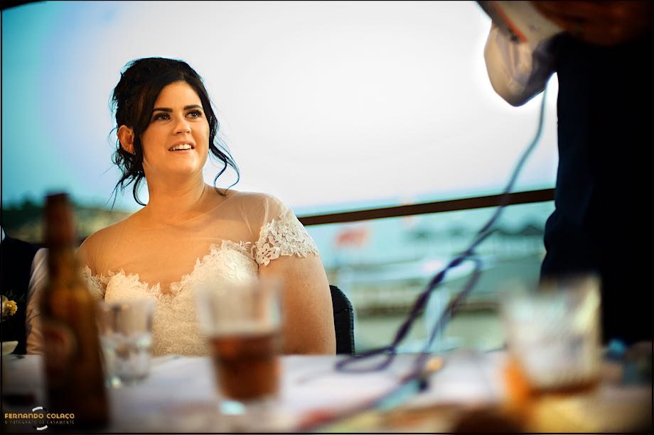 The bride, sitting at the table, smiles as she hears a speech.