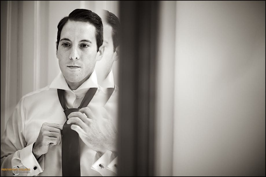 In a mirror on a piece of furniture in his parents' house, the groom tightens his tie.