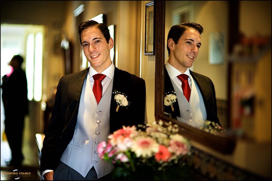 Next to a mirror, on a piece of furniture in his parents' house, which reflects him, the groom looks forward with a happy air.