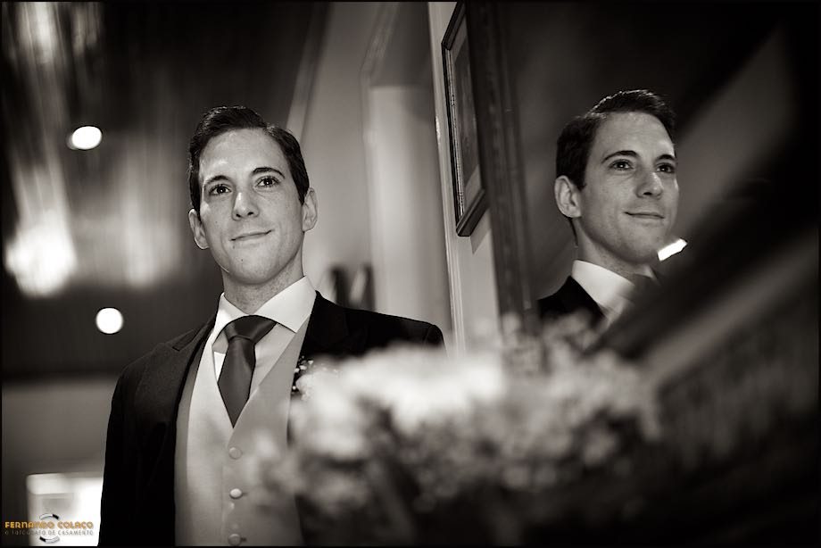 Viewed from below and with a blurred floral arrangement, the groom looks ahead smiling, in a portrait by the wedding photographer, reflected in a mirror.