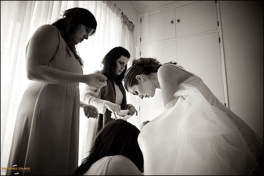 The bride looking at her feet, surrounded by friends who help her put on her shoes, already dressed for the wedding ceremony.