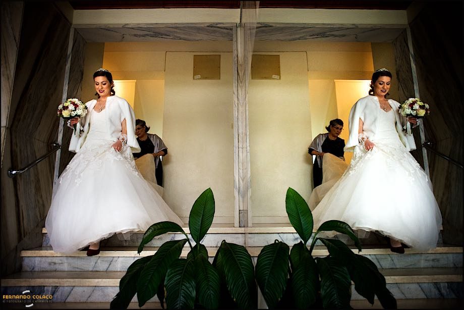 The bride, with her mother helping her with the train of her dress, going down the stairs of the building, where her parents live, reflected in a mirror.