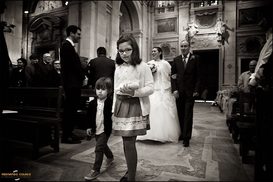 A boy and a girl, with their rings, in front of the bride with her father, walk the path, inside the Basilica of Mafra, to the altar where the groom awaits them.