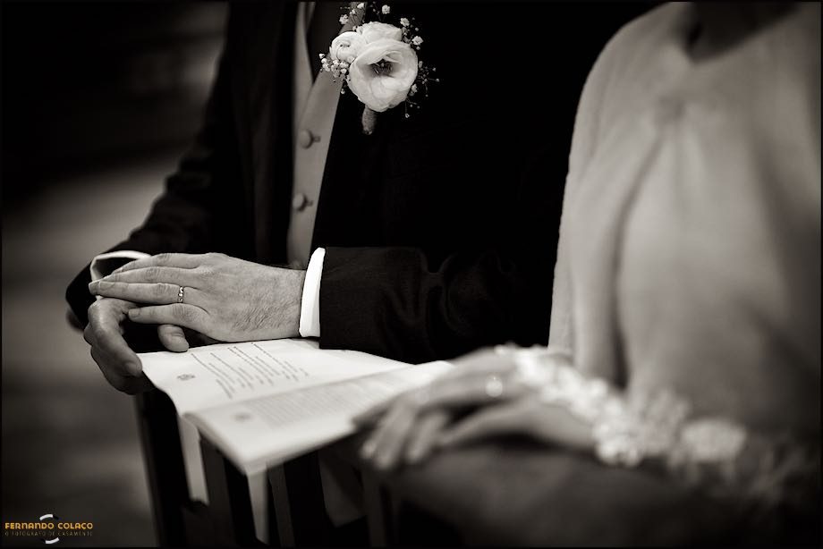 Next to the bride, out of focus, the groom's hands hold the wedding ceremony book, already with the ring and a flower in the lapel of his coat.