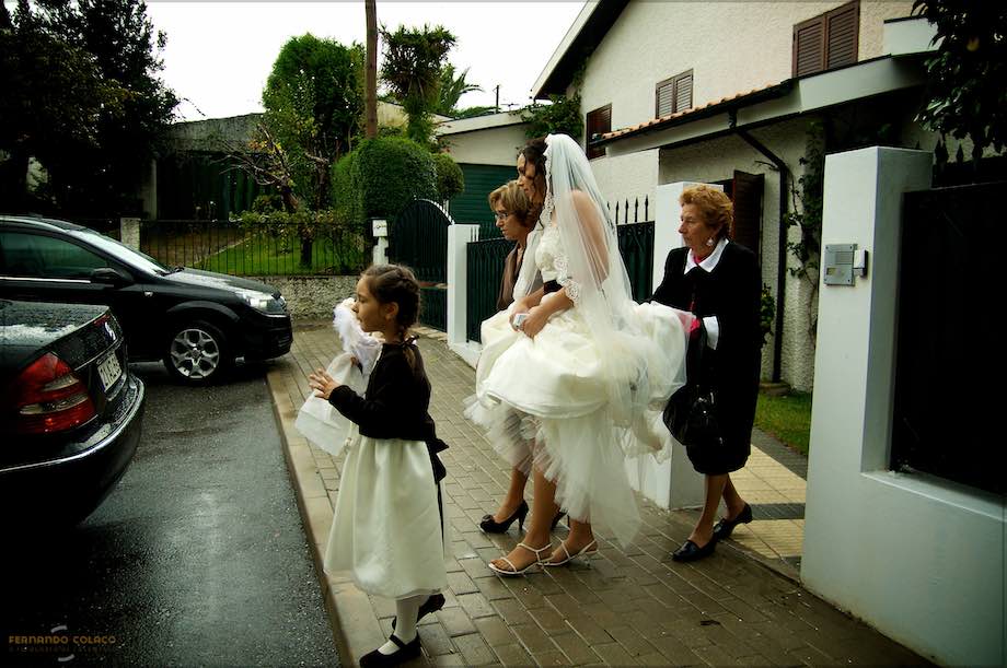With the help of her mother and an aunt, who hold her dress to keep her from getting wet because of the rain, the bride leaves the house to get into the car, along with the girl who is carrying the rings.