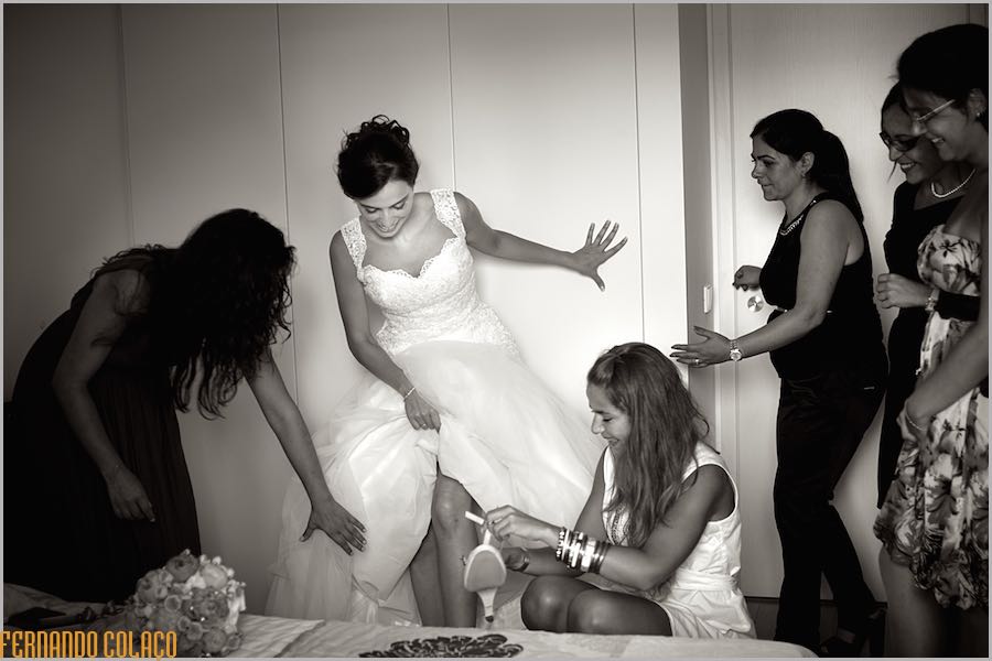 As amigas da noiva, já de vestido, junto dela, quando uma delas lhe calça um sapato, vistas pelo fotógrafo de casamento.