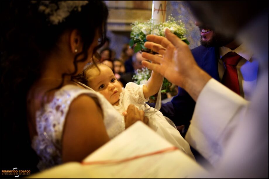 Menina ao colo da mãe a segurar nos dedos do padre, na cerimónia do baptismo.